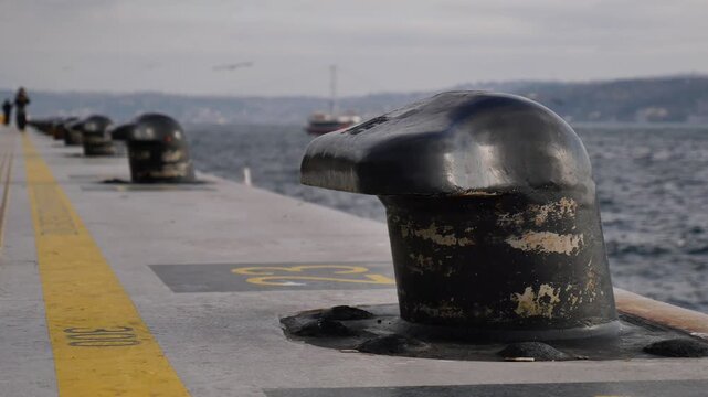 Close up on bollard on pier with seagulls flying over water and distant ships.