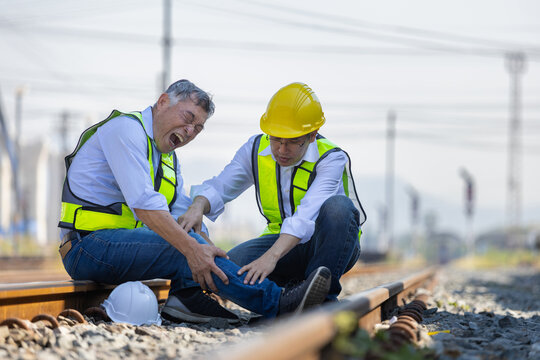 Injured worker having leg pain and coworker helping him at railway construction site, Construction accident with worker suffering from knee injury and colleague assisting outdoors