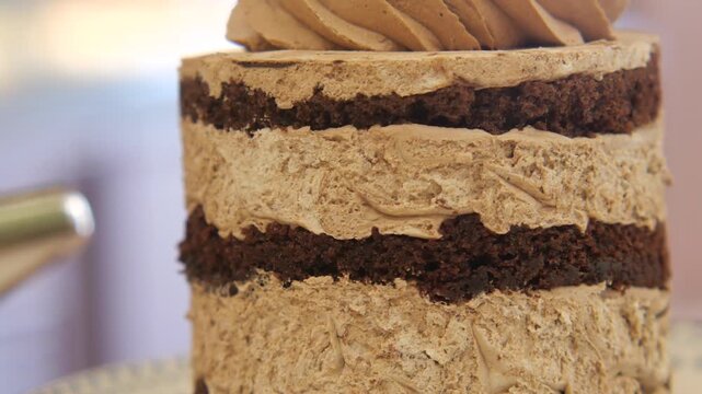 A naked layered chocolate cake with mocha frosting is being decorated with piping bag.