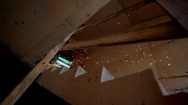 Bright sparks scatter under concrete staircase during metal cutting work. Grinding action sends glowing particles across construction area