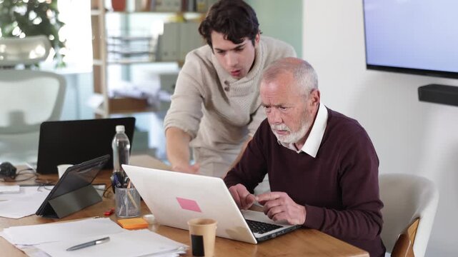 Guy leaned over to seated senior man employee, supervise activities, helps with work and looks at laptop screen. Travel agency, engineering company, notary office or legal advice 