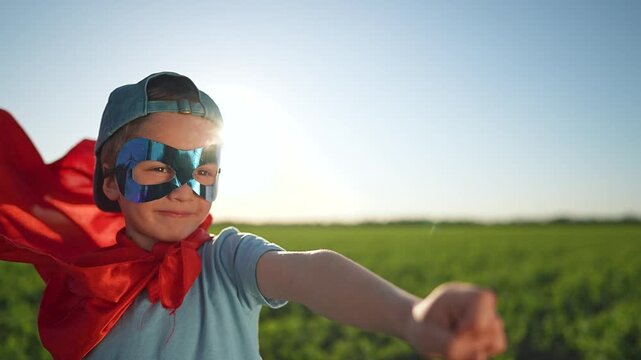 Boy acts like superhero in field, wearing blue mask and red cape. Child raises hand posing like flying. Mask on boy, cape blows in wind. Happy child enjoys outdoor play acting superhero adventure.
