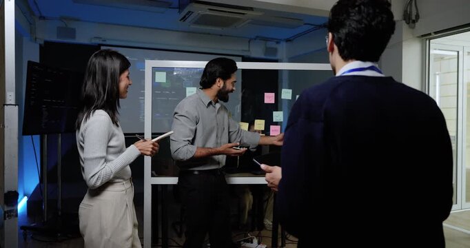 Adult male technology professional gestures toward digital screen and sticky note board while female colleague responds, discussing software workflow during collaborative strategy meeting in office.