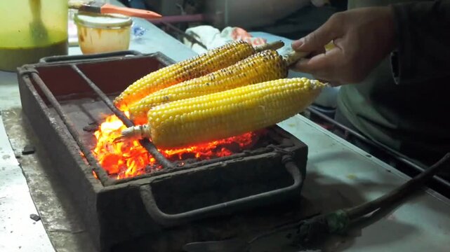 Several fresh corn cobs are grilled over bright charcoal, slowly developing a golden, smoky surface as a vendor carefully tends the fire.