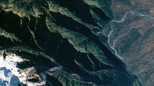 Satellite view of rugged mountain ranges with green slopes, deep shadows, and a winding river flowing through a dry, arid valley landscape below