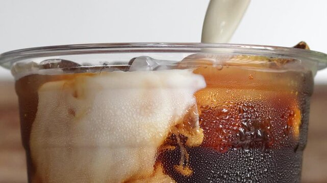 4K footage Close up shot of fresh milk swirling into iced black coffee inside takeaway plastic cup, dynamic fluid motion patterns for iced latte and cold brew coffee menu concept.
