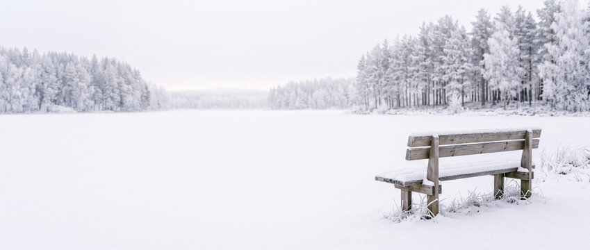 Snow-covered wooden bench by frozen lake in winter landscape  