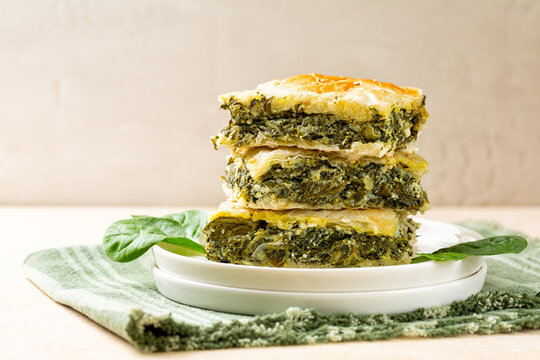 Stack of Spanakopita piece of pie, homemade Greek savory spinach pastry. It contains cheese feta, chopped spinach, green, egg, layered in phyllo or filo dought. Light beige background.