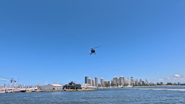 Helicopter flying low over water near city harbor with skyline and boats in summer.