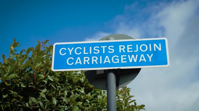 Cyclists rejoin carriageway road sign against blue sky. Blue traffic direction sign on metal post beside green foliage. Cycling route signage with white lettering under partly cloudy daytime sky