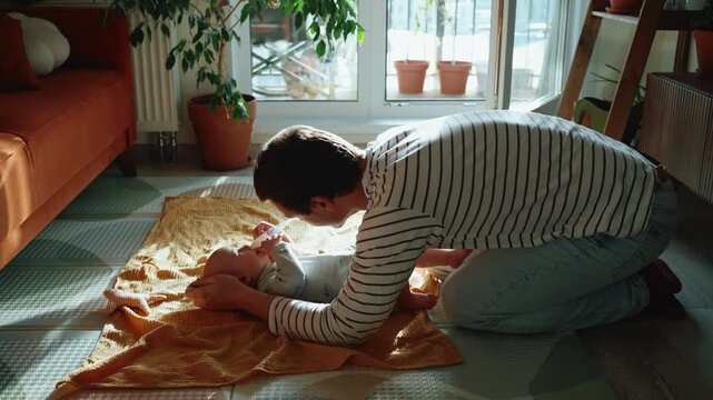 Young dad performing hygiene procedures for his infant child, kneeling on the floor and carefully using a snot sucker to clean the baby's nose from mucus for better breathing