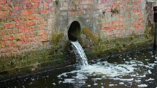 Industrial wastewater pipe discharging into canal water beside brick wall. Foamy polluted runoff below moss covered masonry and drainage outlet. Water contamination from sewer effluent entering urban