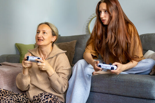 Cheerful mother talking while her disappointed teenage daughter loses a competitive video game. Concept of playful rivalry, gaming competition, and different generational reactions during family play.