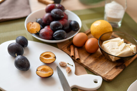 Kitchen table with food ingredients for sweet home seasonal cooking. Sliced plums on cutting board, blurred eggs, butter, sugar and lemon on background