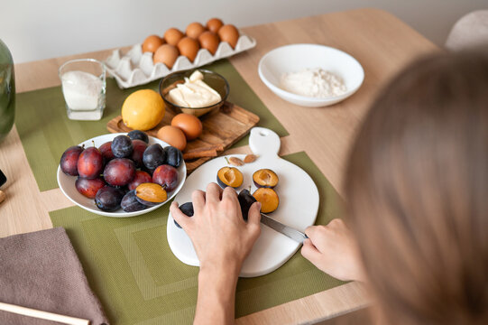 Person cut plums on cutting board, food ingredients for plum pie recipe on table, soft selective focus. Authentic lifestyle home cooking process