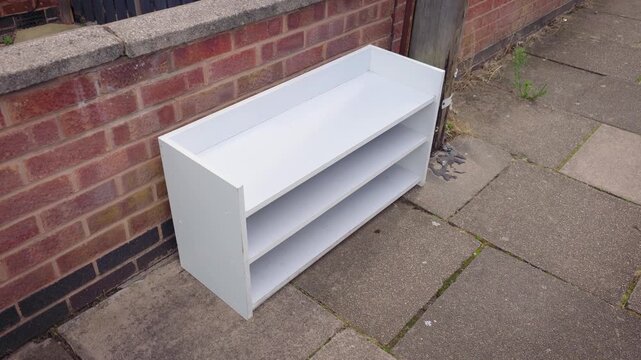 Free shelving unit for collection outside residential property. Used white storage furniture beside brick wall for donation and reuse. Secondhand bookcase available for pickup from sidewalk near home
