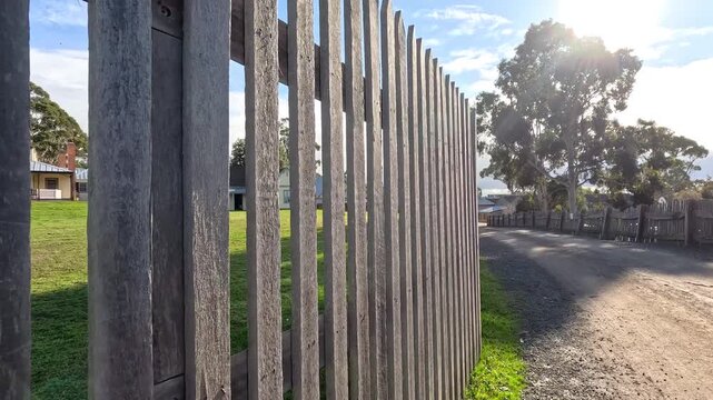 Camera moves slowly along rustic wooden picket fence on sunny day in quiet country neighborhood.