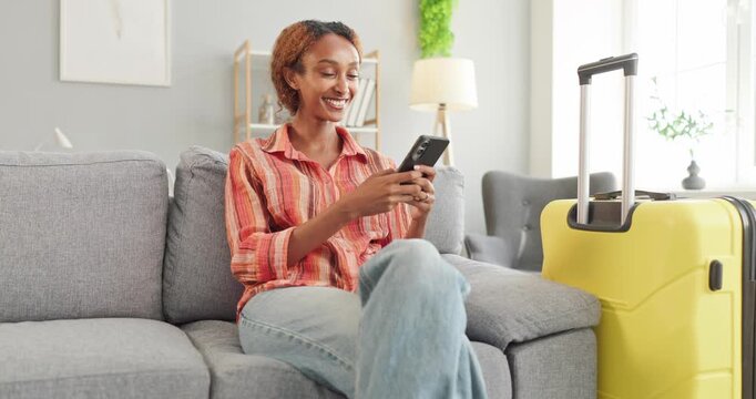 Woman uses smartphone beside suitcase for travel booking. Smiling traveler sits on sofa, arranging vacation details and hotel reservations before departure. Online travel booking made easy.