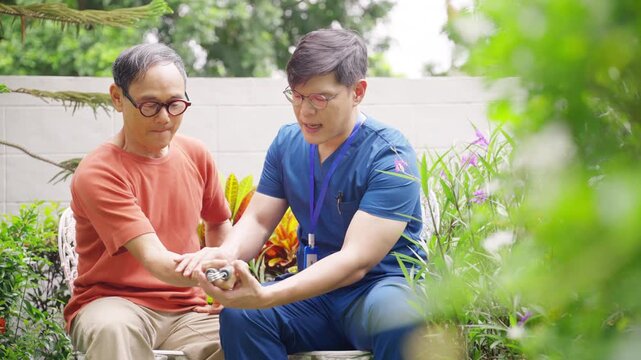 Asian male caregiver or Physiotherapist helping senior man exercise with hand gripper in garden at home, mobility strength training, healthy lifestyle, rehabilitation and elderly wellbeing