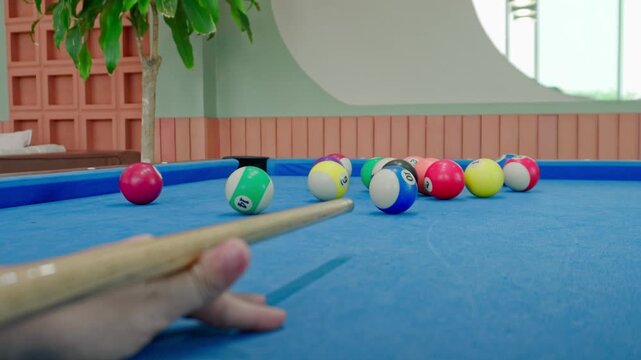 POV shot of person aiming cue at pool balls on blue felt. Focused player carefully lining up a deliberate break shot on a vibrant blue table in a cozy retro lounge with plants and seating.
