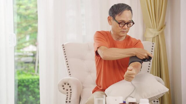 Asian senior man measuring blood pressure at home using digital monitor, elderly person checking hypertension and cardiovascular health during daily healthcare routine and wellness monitoring