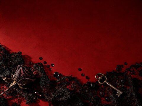 A dried rose and an antique metal key resting on a dark black lace fabric against a deep red textured background, creating a mysterious and romantic gothic atmosphere