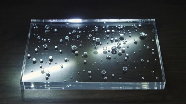 Camera recording acrylic block, droplets merging, forming beads on dark wood under overhead light