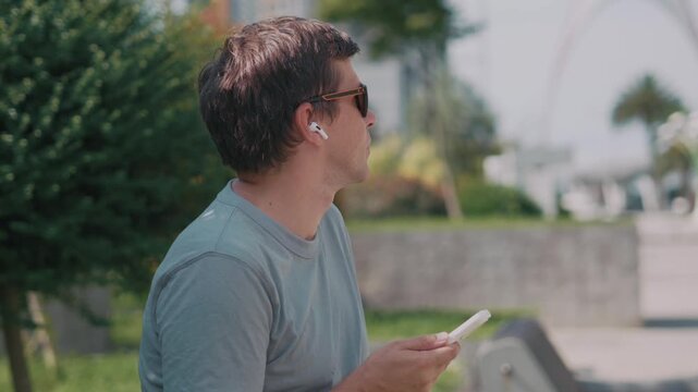 Man using smartphone with wireless earbuds outdoors, choosing music or podcast and listening to digital audio during relaxed break in city park.