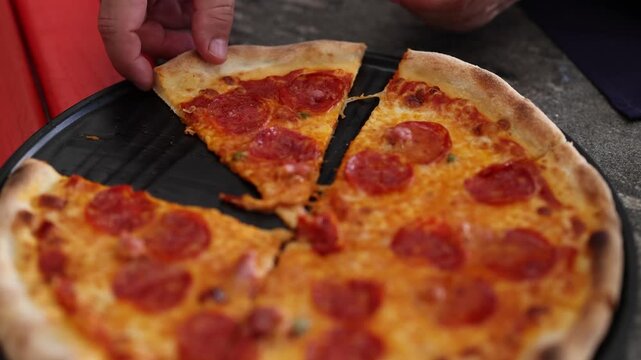 Close up cinematic shot of person taking a slice of hot delicious pepperoni pizza with melted cheese from a round black pan.