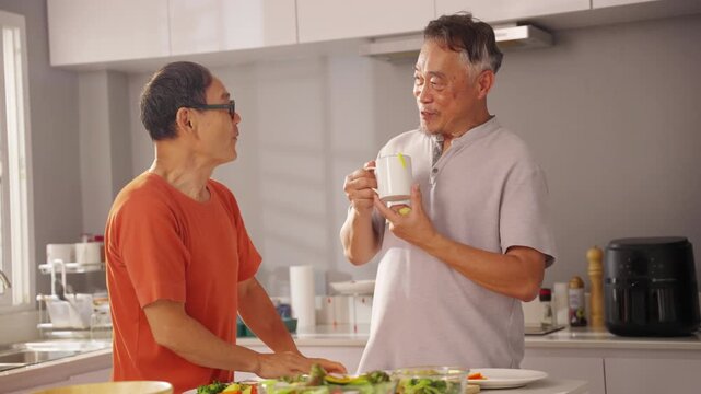 Happy Asian senior gay man couple preparing and enjoying healthy organic dinner together in kitchen at home, cooking fresh vegetables and sharing wellness routine for active retirement lifestyle.