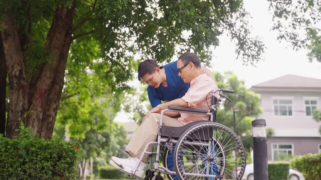Asian male caregiver helping senior man stand up from wheelchair  during outdoor walk near home. Elderly nursing health care, mobility support and mental wellbeing. Retirement home healthcare.