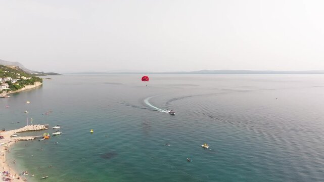 Red parasail and tow boat cruise past a crowded Croatian swimming cove as swimmers and inflatables dot the wide open sea.