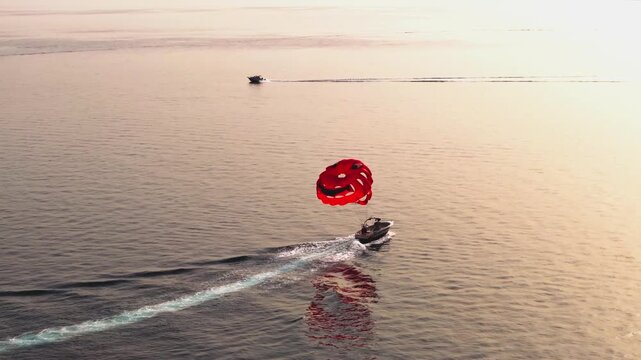 Red parasail and tow boat cruise across soft golden water as another powerboat crosses the frame in the distance.