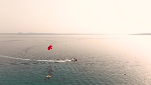 Red parasail and tow boat arc over quiet open water as smaller craft float nearby on a soft hazy day.
