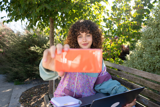White woman taking selfie on bench outdoors with laptop and notebook