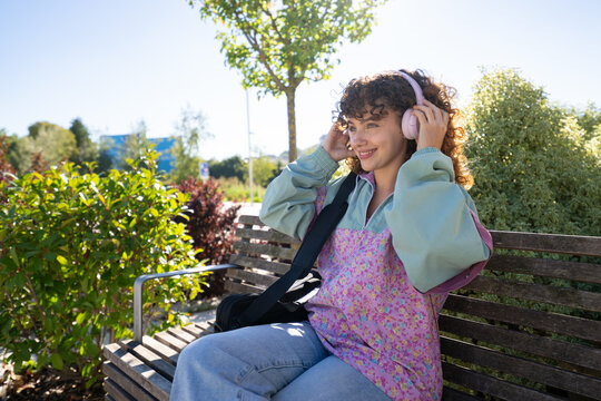 White woman sitting on bench wearing headphones in sunny park