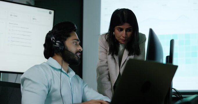 Adult male technology professional wearing headset points at computer screen while female colleague leans beside workstation, discussing software information during technical support in office.