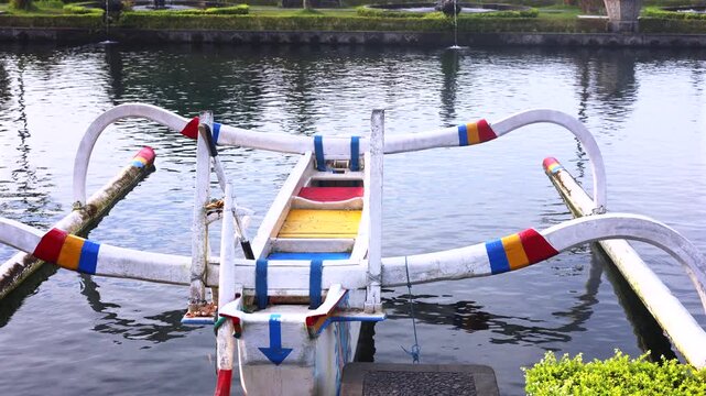 Traditional outrigger canoe floats in still river water, with painted support arms and narrow hull centered in frame. Boat remains docked near landscaped bank, suggesting peaceful transport.