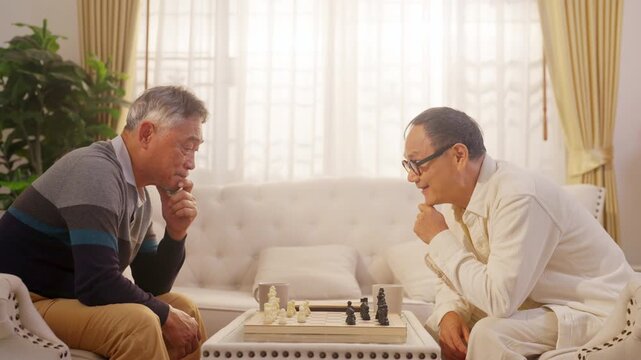 Happy Asian senior gay couple playing chess together at home. Elderly family enjoying brain exercise leisure activity strategic board game, mental wellbeing during relaxed retirement active lifestyle