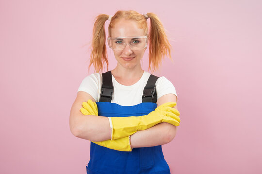 Confident young woman wearing blue overalls, yellow rubber gloves, and safety glasses, standing with arms crossed against a solid pink background, ready for demanding tasks