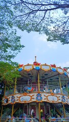 Ornate two-tier carousel ride surrounded by green trees under clear sky  © IvanRheza