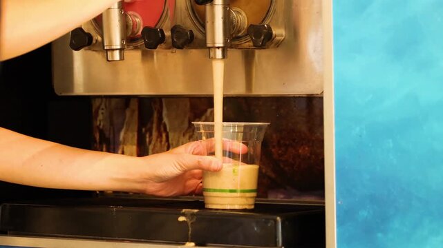 Close up of hands pouring cold yellow slushy drink from dispenser machine into plastic cup in snack shop.