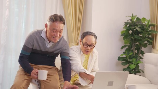 Happy Asian senior gay couple drinking coffee while reviewing income, expenses and bank statement together on laptop at home for household budgeting, financial planning and retirement money management