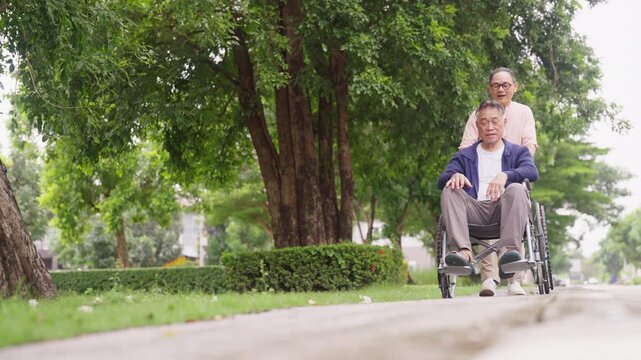 Asian senior gay couple caring for each other during outdoor relaxing together in public park. Elderly man push partner in wheelchair walking on footpath, caregiving mobility support healthy wellness