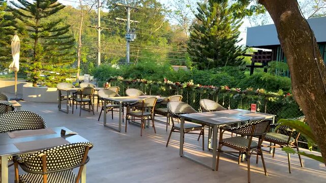 A serene empty outdoor cafe patio with tables and chairs surrounded by lush green trees and natural light, inviting relaxation.