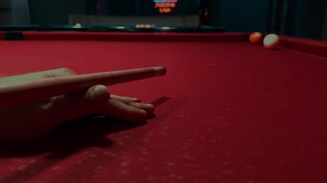 A player strikes a white cue ball across a red pool table with a wooden cue. The close view shows the hand position and rolling balls during the game.