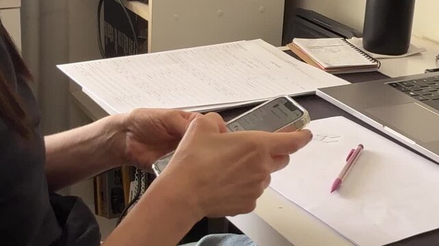 Scrolling and using a smartphone while seated at an office desk with laptop, paperwork and notebook, illustrating digital communication and everyday work, handheld close-up