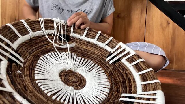 Time lapse video of a woman wearing glasses quickly weaving white macrame cord onto a natural banana bark wall decor piece indoors.