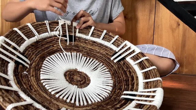 Slow motion video of a woman's hands skillfully weaving and stitching white macrame yarn onto a natural banana bark wall decor piece indoors.
