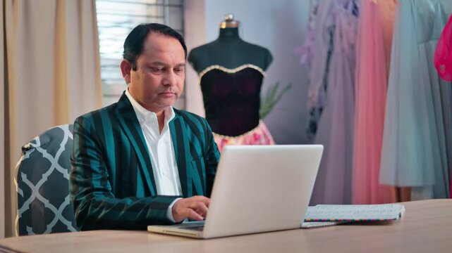 Senior Indian male fashion designer working on a laptop in a creative studio surrounded by dresses and a mannequin.